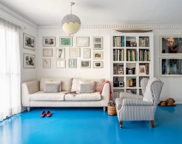 Foto mostra sala de estar com piso azul vibrante, sofá e poltrona em tons neutros e parede com arranjo de quadros e estante de livros.