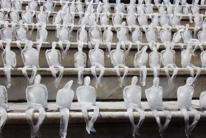 Pequenas esculturas de gelo derretendo de pessoas sentadas uma ao lado da outra de cabeças levemente baixas numa escadaria.