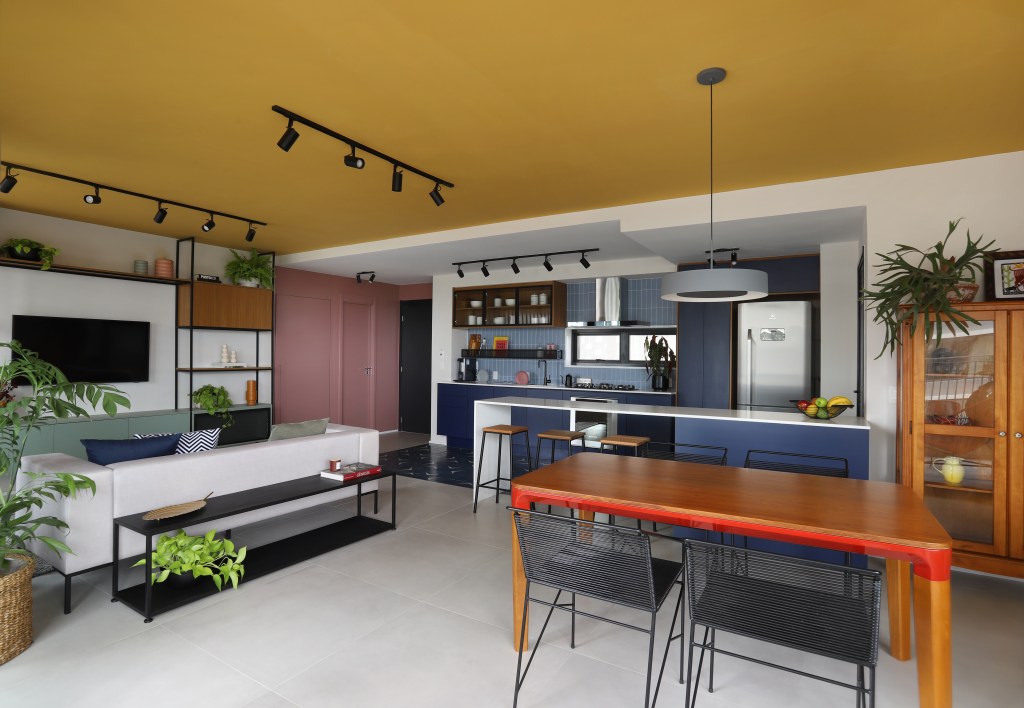 Sala de estar integrada com jantar e cozinha; teto amarelo; mesa em madeira; sofá cinza; hall rosa; cozinha com marcenaria azul