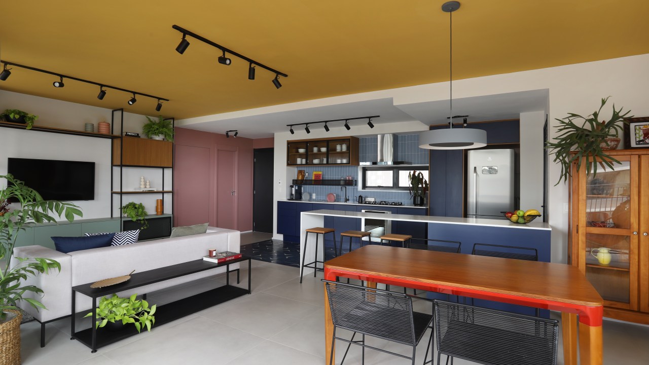 Sala de estar integrada com jantar e cozinha; teto amarelo; mesa em madeira; sofá cinza; hall rosa; cozinha com marcenaria azul