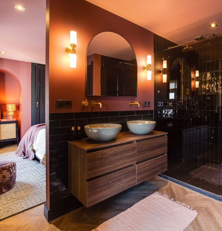 Banheiro com pia dupla sobre uma bancada de madeira escura. Azulejos pretos fazem o revestimento do box e da parte de fora até a altura do espelho. A partir daí a parede é pintada em um tom de rosa.