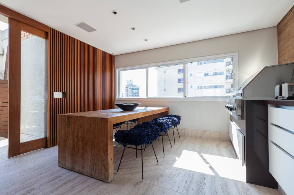 Foto mostra cozinha com mesa de madeira e banquetas em tecido azul marinho. Na lateral esquerda vê-se um painel ripado de madeira que 'esconde' a porta de correr, também de madeira.