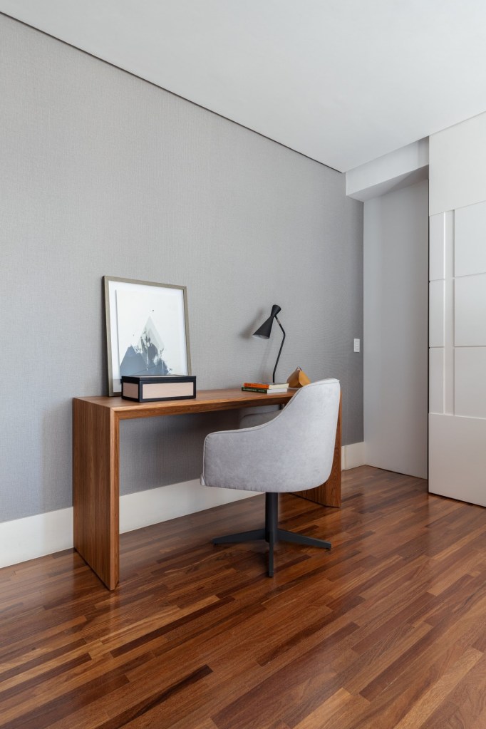 Foto mostra home office com mesa de madeira e cadeira giratória com acabamento em tom de cinza. O rodapé é branco, assim como a porta de acesso ao ambiente. O piso é de madeira.