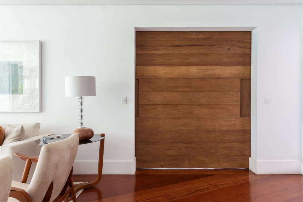 Foto mostra porta de madeira em parede branca, com rodapé na cor branca e piso de madeira. Há mesa lateral com abajur em tons neutros.