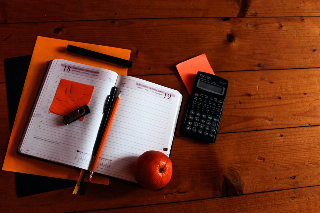 Foto mostra agenda aberta sobre tampo de mesa de madeira, com lápis, caneta, pendrive, calculadora ao lado e post its na cor laranja. E tem uma fruta ao lado também.