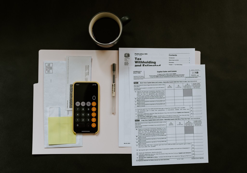 Foto mostra papeis sobre mesa com calculadora por cima e xícara de café logo acima, sobre a mesa.