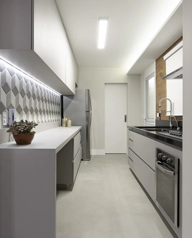 Foto mostra cozinha com bancada em que se vê fita led logo abaixo dos armários aéreos, iluminando área de trabalho e preparo dos alimentos.
