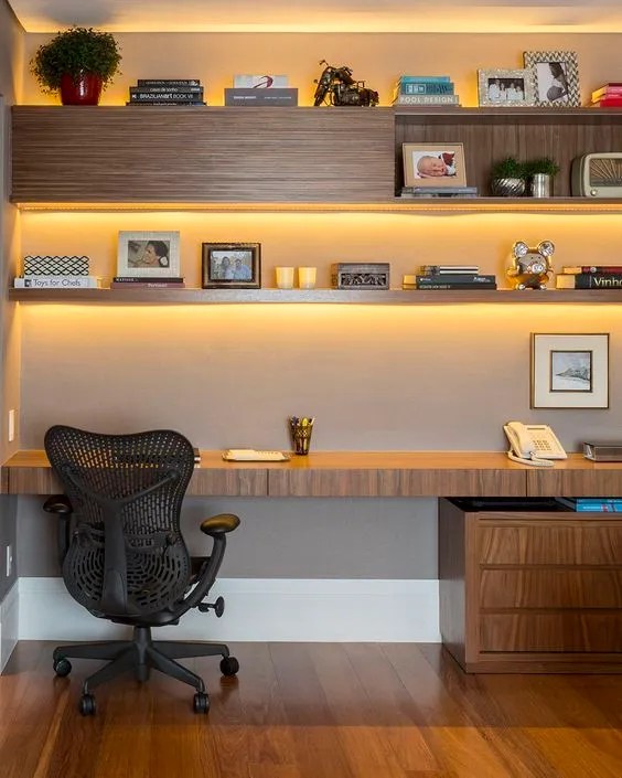 Home office tem mesa de madeira com prateleiras do mesmo material, que receberam fitas led para iluminar melhor cada detalhe.