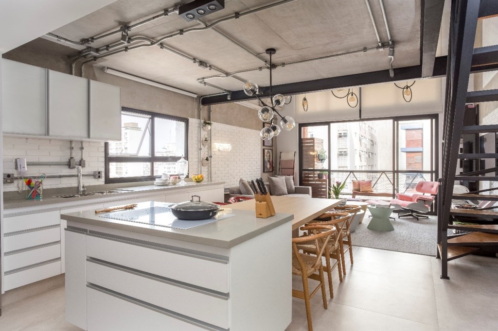 Foto mostra cozinha integrada à sala de estar com tubulação aparente em tom prateado, combinando com paleta de cores neutras e tons claros.