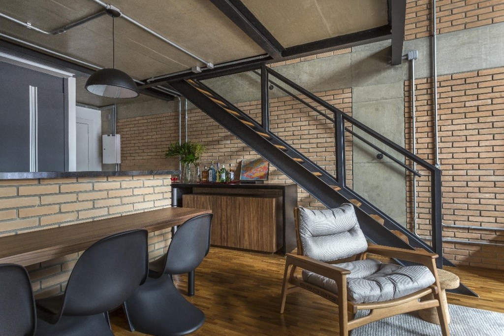 Foto mostra sala de estar com escada e tubulação aparente compondo o visual em estilo industrial.