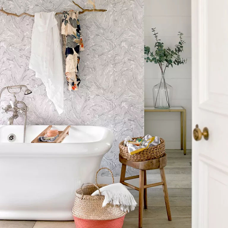 Banheiro com uma banheira, um banco de madeira, um cesto e uma parede com papel ilustrado.