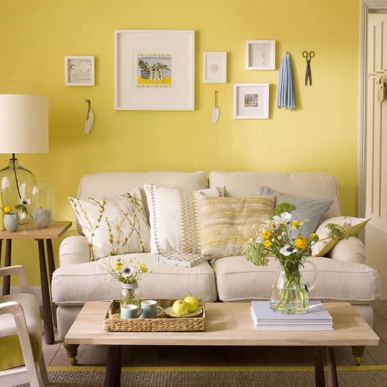 Sofa creme, parede amarela, mesa de centro com tampo de madeira, alguns quadros pendurados e uma mesa lateral com um abajur.
