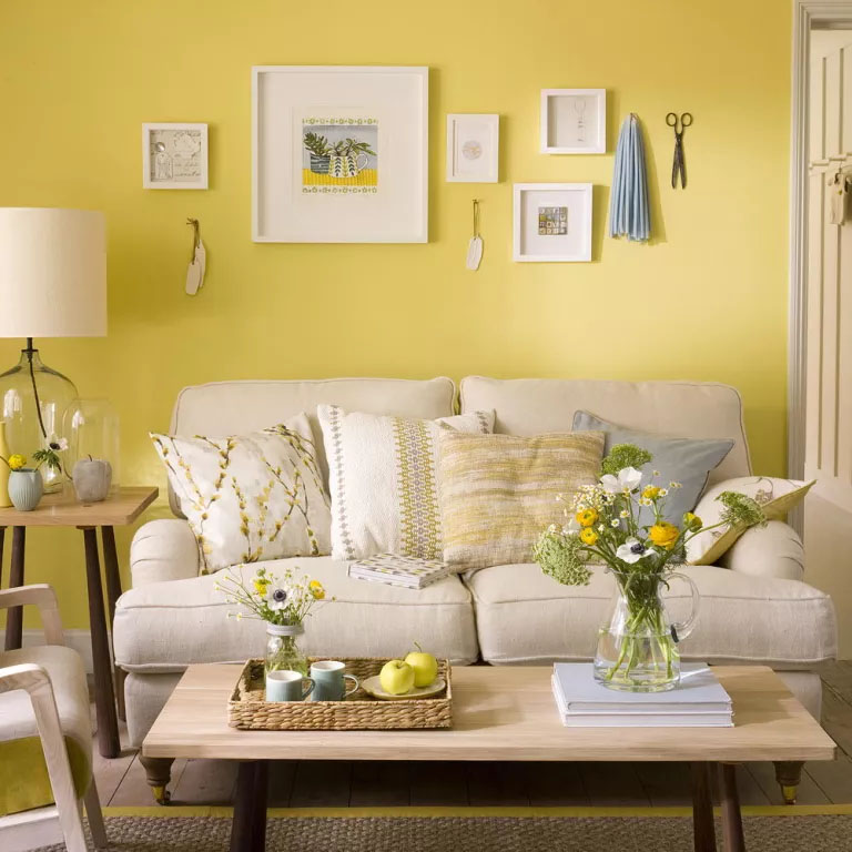 ideias-parede-sofa Sofa creme, parede amarela, mesa de centro com tampo de madeira, alguns quadros pendurados e uma mesa lateral com um abajur.