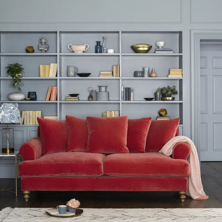 Sofá de veludo vermelho. Ao fundo, um grande armário com diversas prateleiras, pintado de cinza.