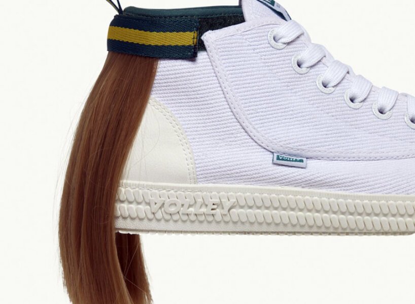 Tênis todo branco com uma mecha de cabelo presa na parte superior traseira, simulando um mullet. O cabelo é preso por uma tira de velcro que por fora é verde com uma listra amarela.