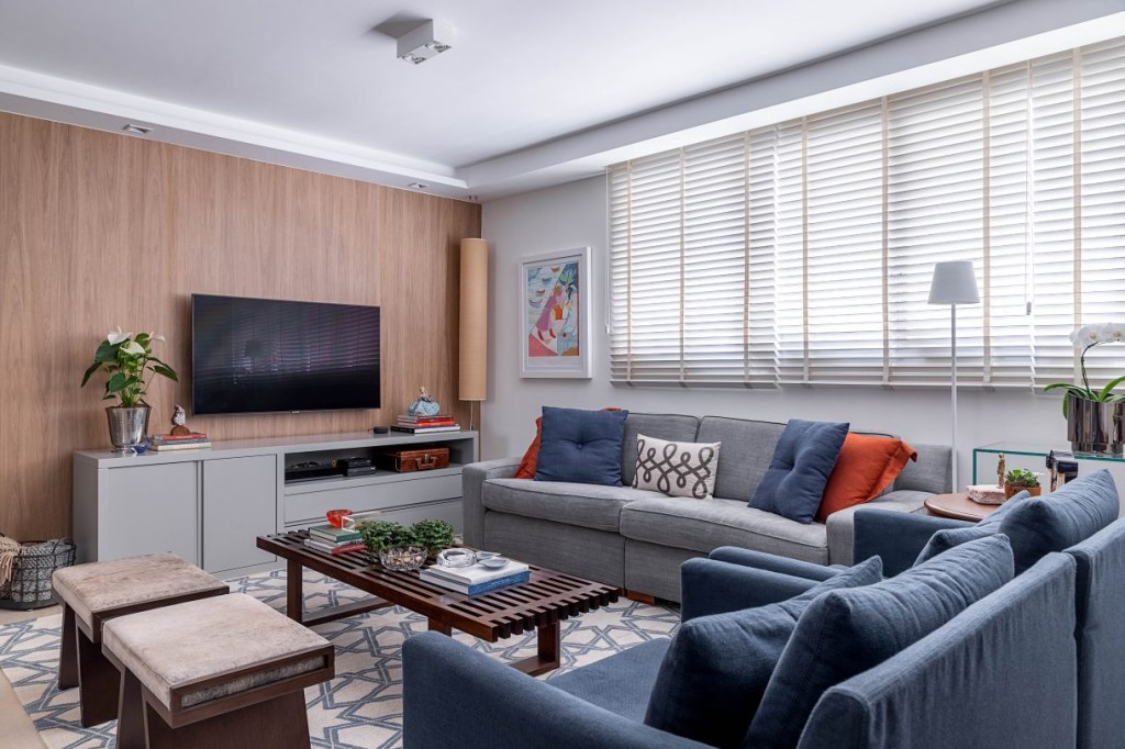 Visão angular da sala, dois sofás, um azul e outro cinza, grande tapete branco com detalhes geométricos em azul, televisão presa a um painel de madeira
