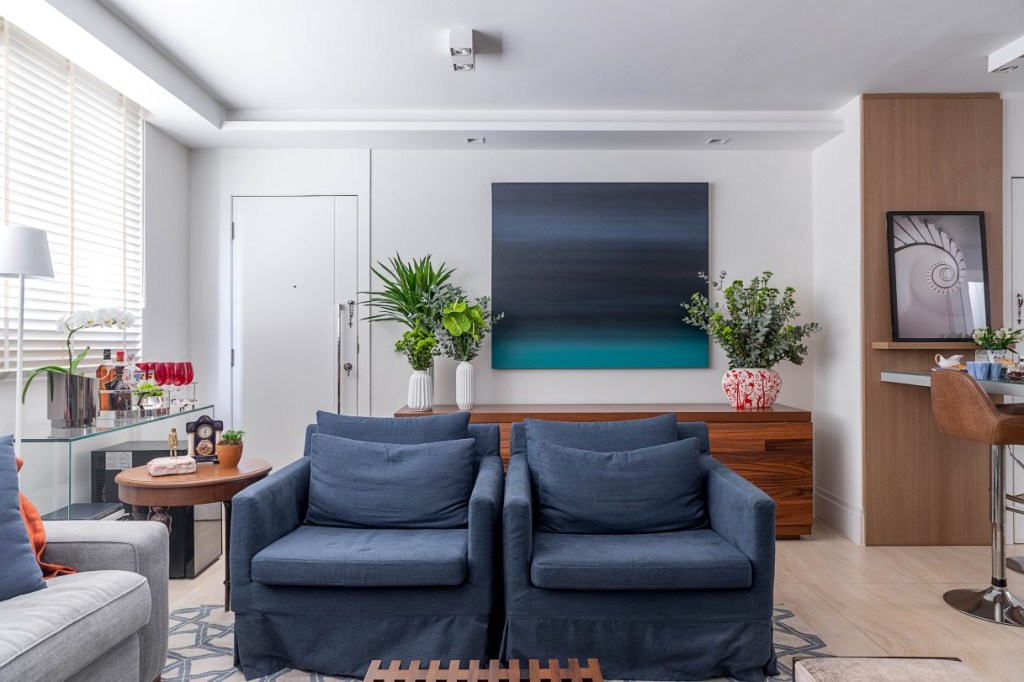 Sala pequena com sofá azul virado para um grande quadro com diferentes tons de azul, janela na lateral esquerda e paredes brancas.