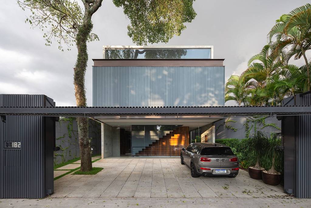 Fachada da frente tem portão metálico da garagem levantado, onde se vê a integração do espaço do térreo com o jardim da casa.