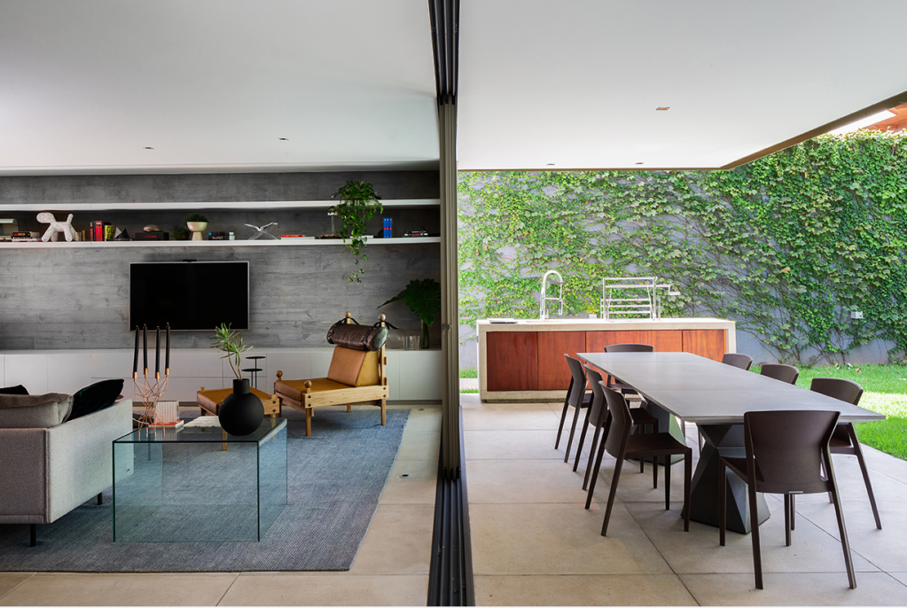 Foto mostra integração, por esquadrias metálicas, da sala de estar com a varanda e o jardim, onde vemos uma mesa de jantar e uma bancada de trabalho no estilo cozinha gourmet.