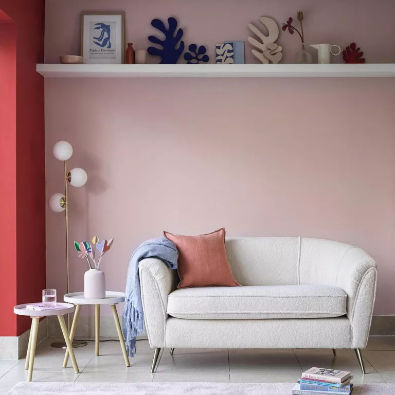 Sofá branco em frente a uma parede rosa, que tem uma prateleira fixa em uma posição muito acima do sofá.