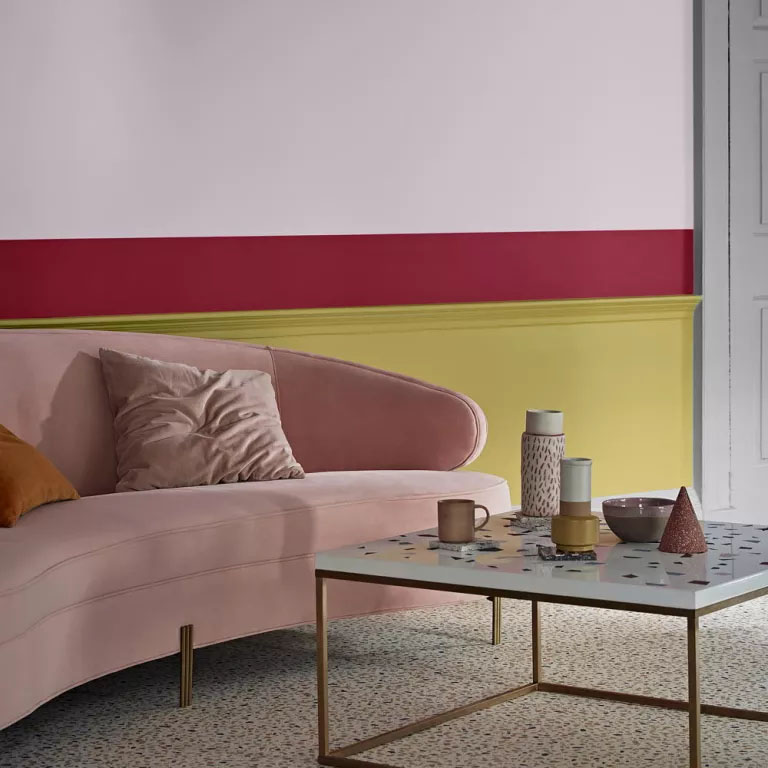 Sofá rosa, uma mesa de centro quadrada, uma parede pintada em três cores: amarelo, vermelho e rosa.