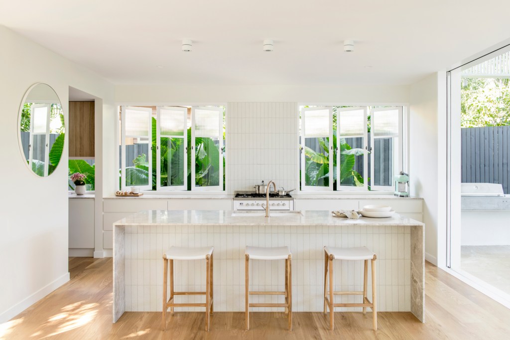 Foto mostra cozinha com bancada branca e três banquetas em madeira e assento branco. área de trabalho ao fundo, com janelas de vidro que deixam o verde 'entrar' no ambiente.