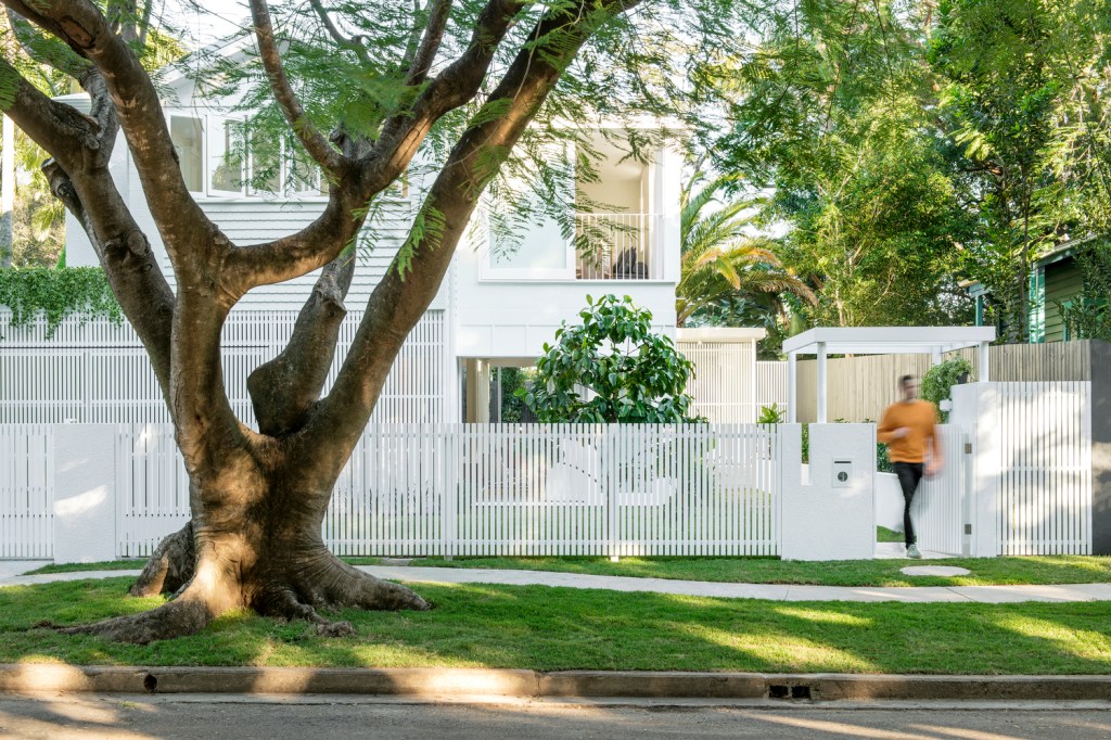 Foto mostra fachada frontal da casa, toda branca, com muito verde e homem saindo pelo portão de entrada. Em primeiro plano, há uma grande árvore na calçada.