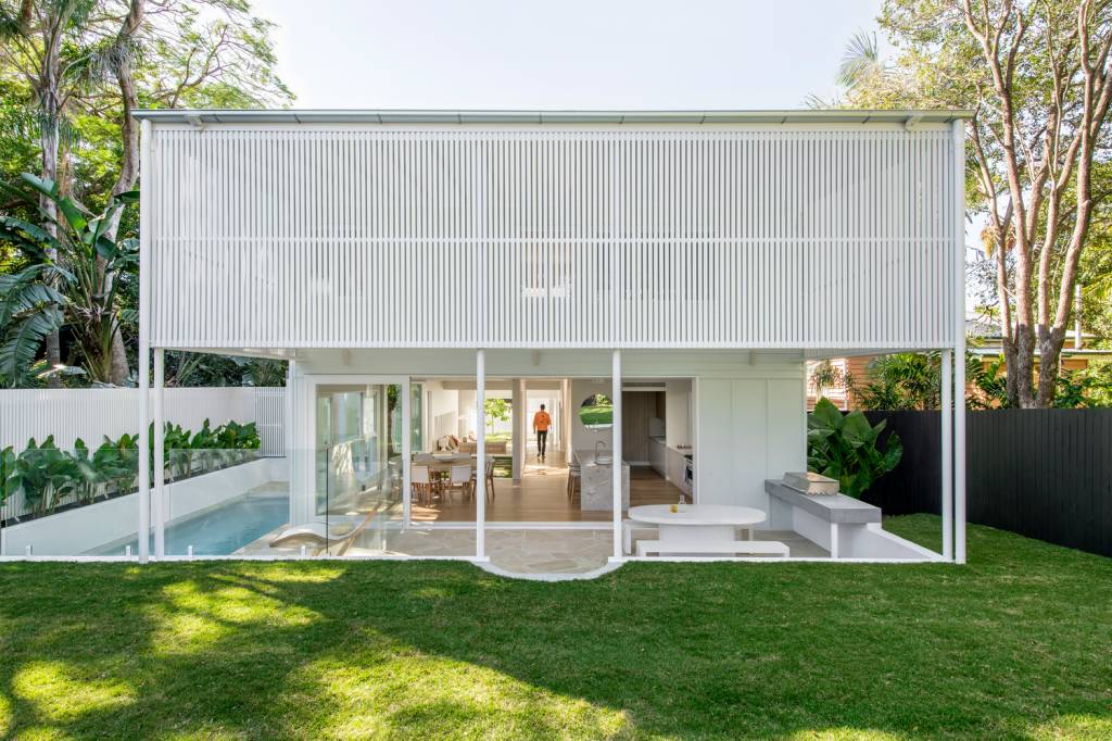 Foto mostra fachada dos fundos de uma casa branca, com gramado em primeiro plano, piscina em raia na lateral, estar integrado no térreo e piso superior com brises.