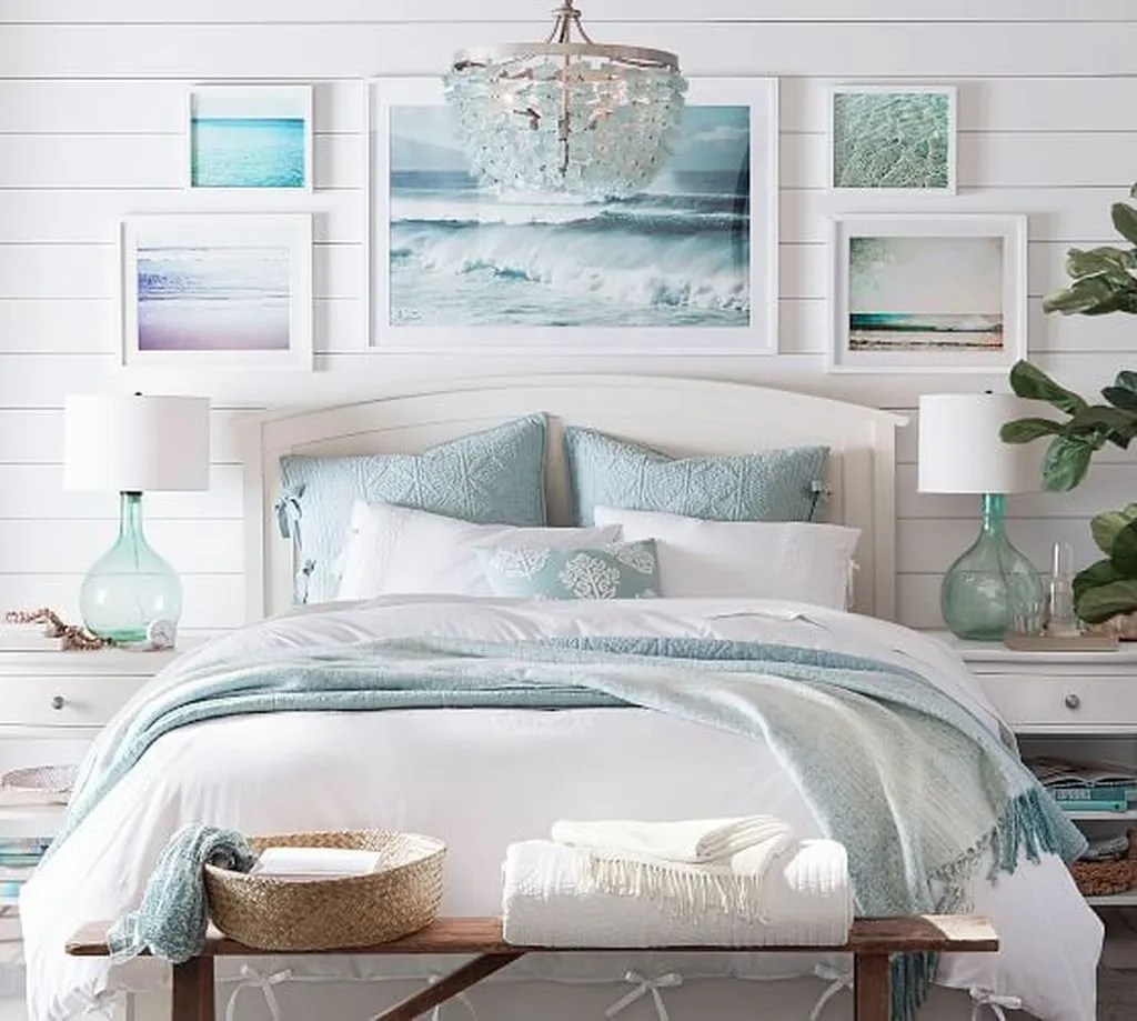 Quarto com cama de casal e roupas de cama brancas e azuis claras; quadros com imagens de mar na parede