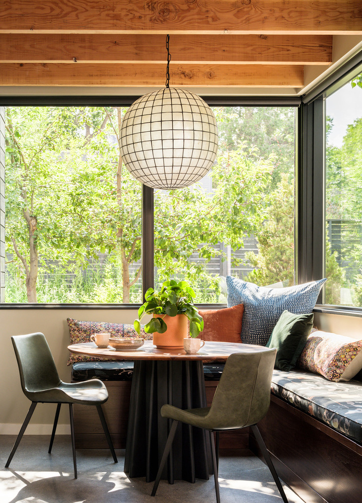 Foto mostra canto da sala de estar com mesa redonda e duas cadeiras. Ao redor, um banco de alvenaria oferece espaço para mais pessoas. A janela de vidro conecta o ambiente ao jardim externo.