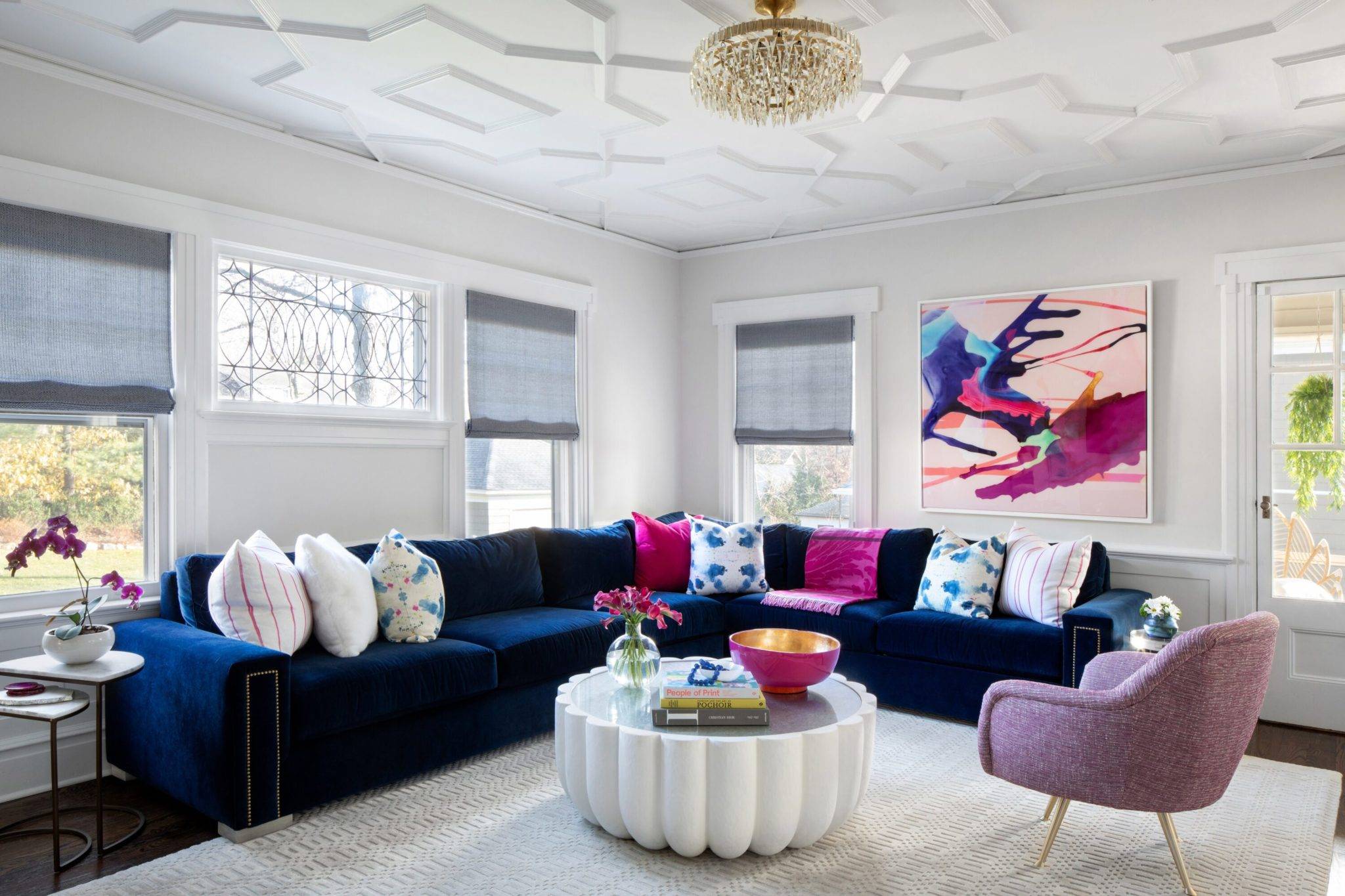 Ampla sala de estar com poltrona rosa, mesa de centro branca, paredes brancas, cortinas em azul claro, e um grande sofá em L azul veludo.