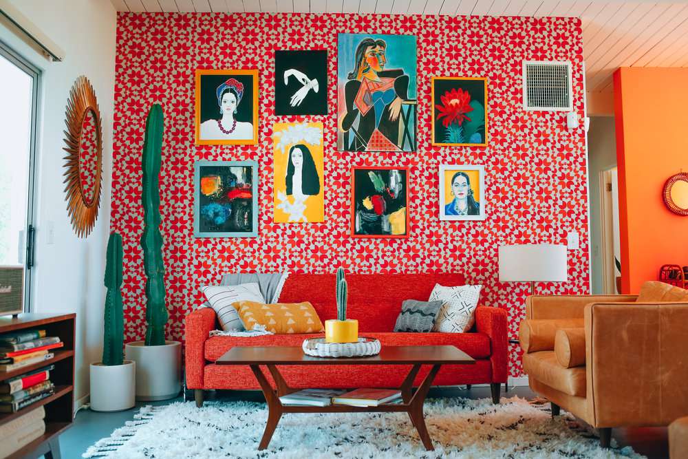 Sofá vermelho, parede com papel de parede com figuras geométricas e diversos quadros pendurados.