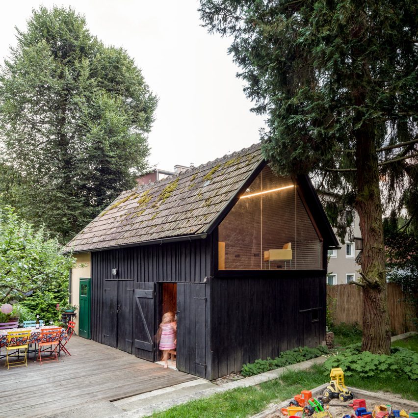 Galpão de madeira em tom de preto, com telhas marrons, tem porta de madeira com uma criança e, no piso superior, uma abertura triangular de vidro.