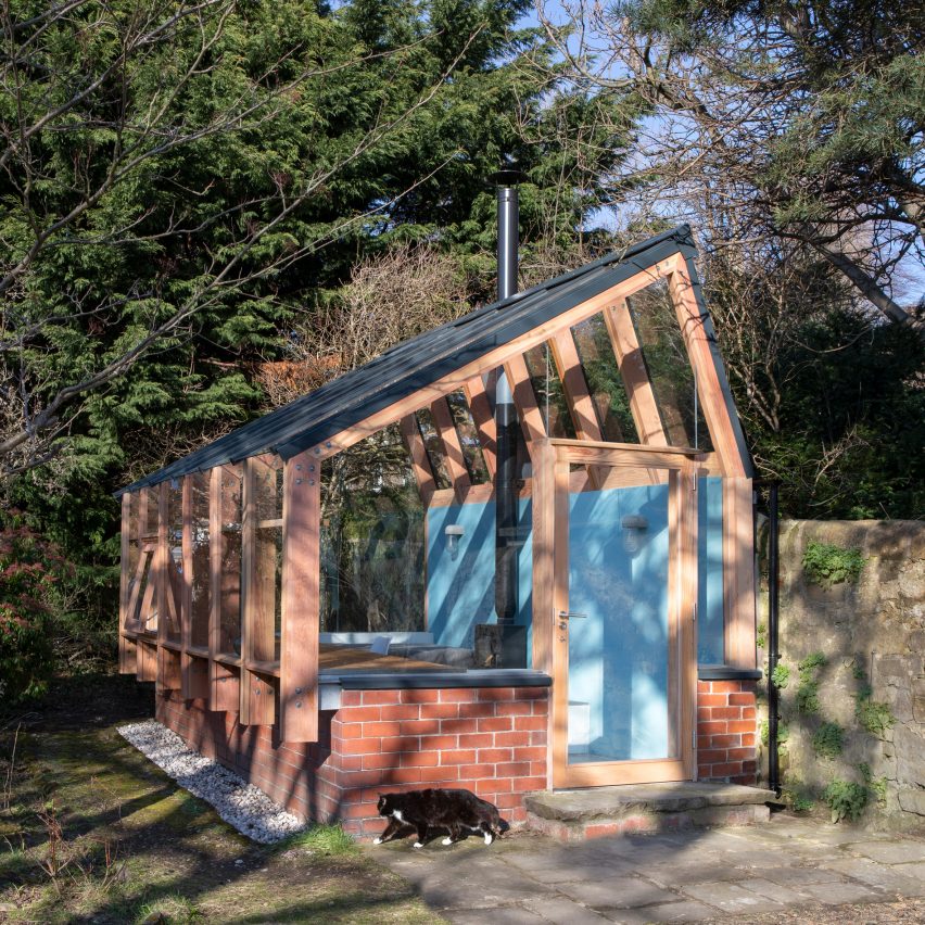 Fachada lateral com porta em que se vê base de tijolos, estrutura de madeira e telhas translúcidas, em um jardim.