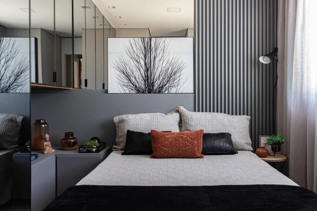 Quarto com cama de casal em primeiro plano, armários com espelho na lateral esquerda, um quadro com árvore de galhos secos na cabeceira e parede em tom de cinza.