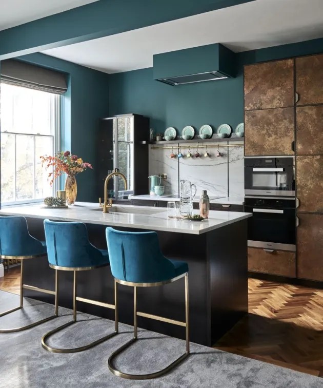 Bancada de cozinha com três banquetas em estrutura dourada e revestimento em tecido aveludado azul, voltadas para a área de trabalho da cozinha, com parede em tom de verde escuro.