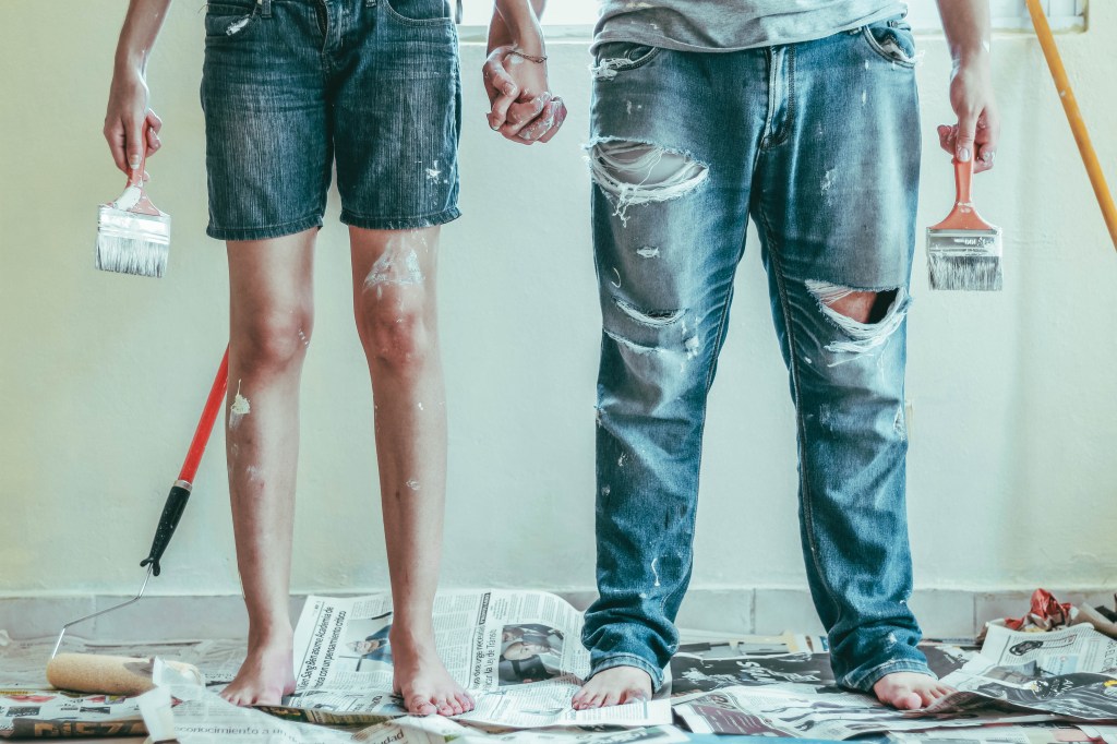 Foto mostra uma mulher e um homem de mãos dadas, apenas da cintura para baixo, em cena que nos leva a pensar que estavam pintando uma parede. Tem resíduos de tinta no corpo deles e eles estão pisando descalços sobre jornais que protegem o piso.