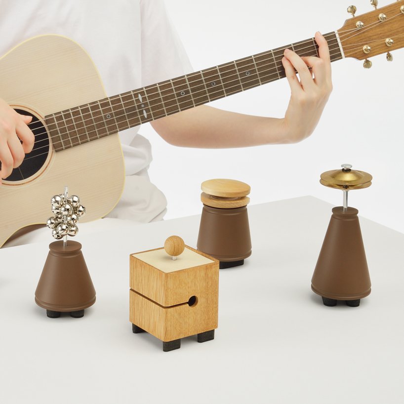 Pessoa toca violão em frente a mesa sobre a qual jazem 4 pequenos objetos de madeira de diversos formatos.