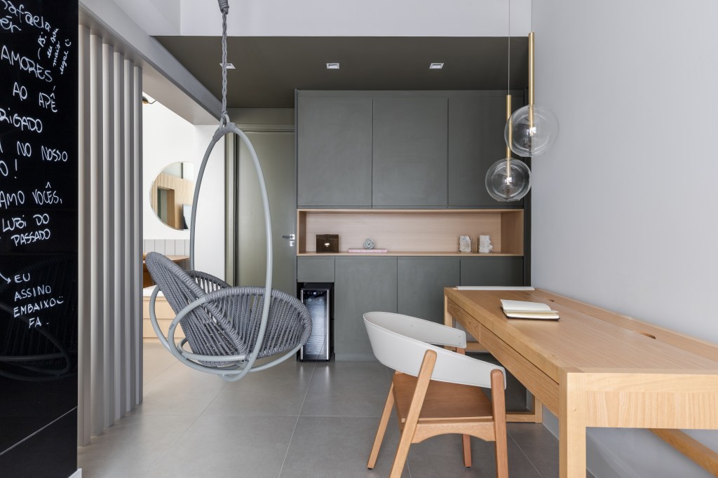 Cadeira suspensa faz uma separação entre espaços de uma sala integrada a cozinha, o balanço foi instalado para criar um ambientação ideal para trabalhar e relaxar.