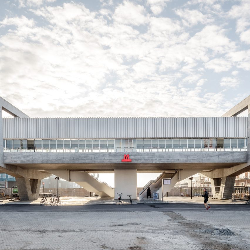 Estação de metrô acima do solo e suspensa por estrutura de concreto. Ao fundo, céu azul com nuvens e sol.