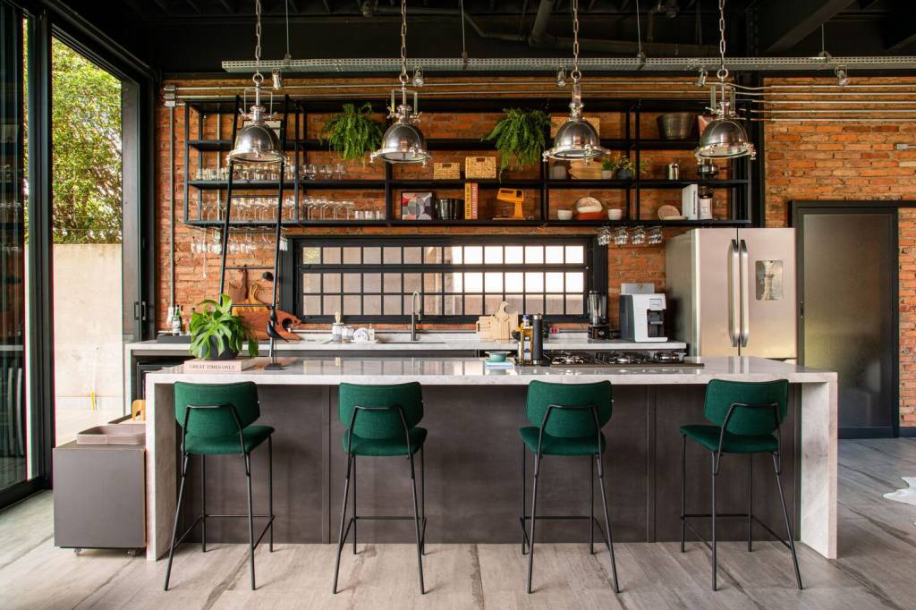 Bancada de cozinha com banquetas em metal preto e estofado em tecido verde. Parede do fundo com tijolinhos e pendentes e prateleiras metálicos em estilo industrial.