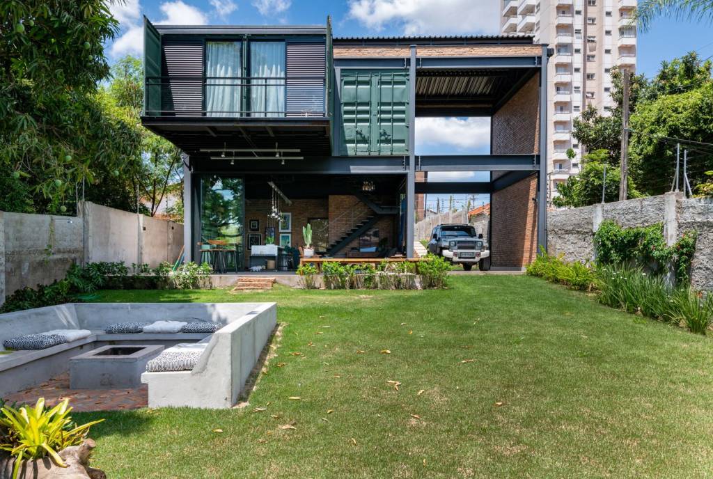 Fachada externa de casa mostra contêiner verde com parte em balanço formando uma pequena varanda, sobre piso térreo com revestimento em tijolo aparente.