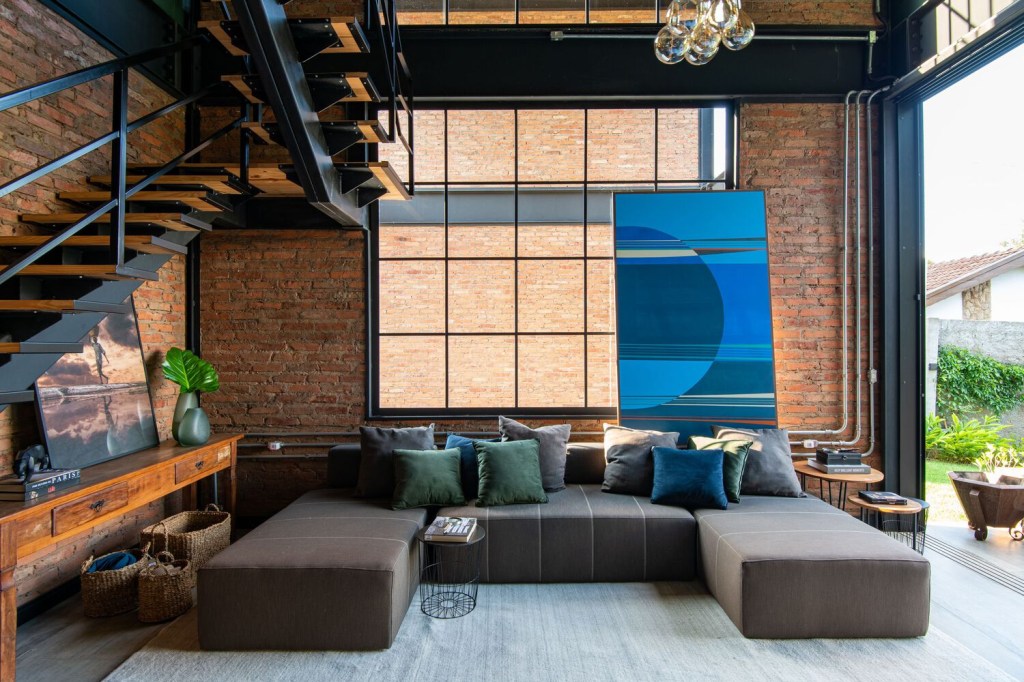Sala de estar com sofá modular em "U", almofadas, uma escada metálica na lateral esquerda e, ao fundo, grande abertura em vidros quadrados, com uma tela de tons azuis pontuando cor.