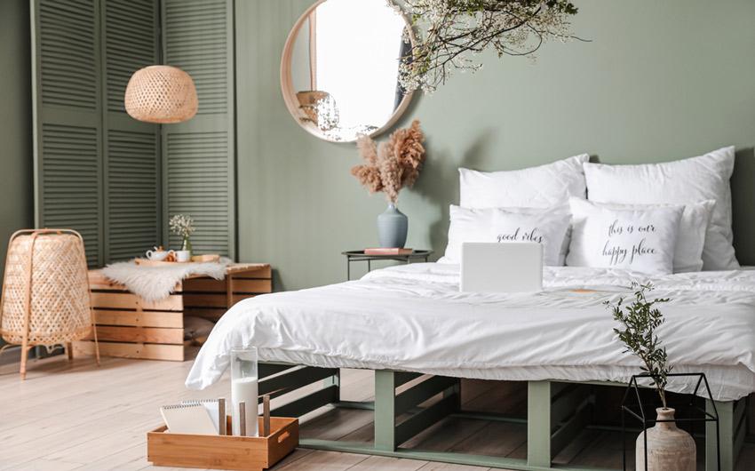 Quarto com cama de caixotes, paredes verdes, espelho circular e arranjos de plantas secas