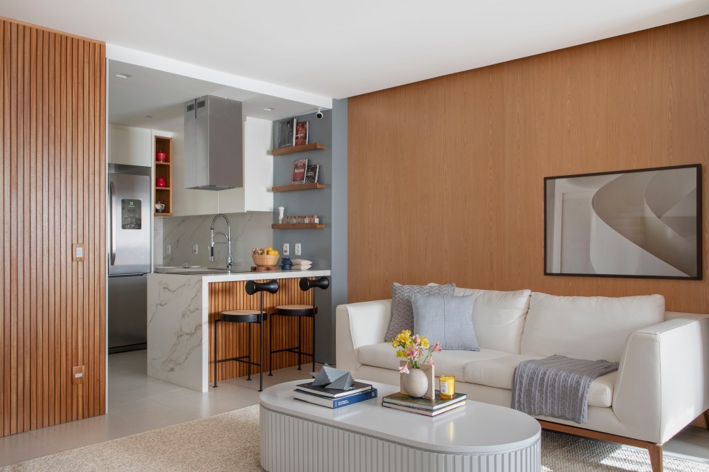 Sala de estar com painel de madeira ripado, sofá branco, mesa de centro oval, integrada com a cozinha de bancada de pedra