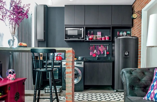 Foto mostra cozinha com armários revestidos com laminado preto fosco, bancada de refeição com tampo de concreto e estrutura de tijolos aparentes.