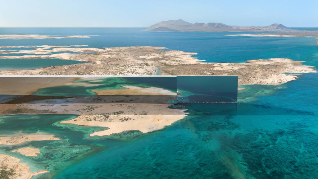 Prédios em forma de muralhas correm paralelos em região desértica a costa de um mar azul.