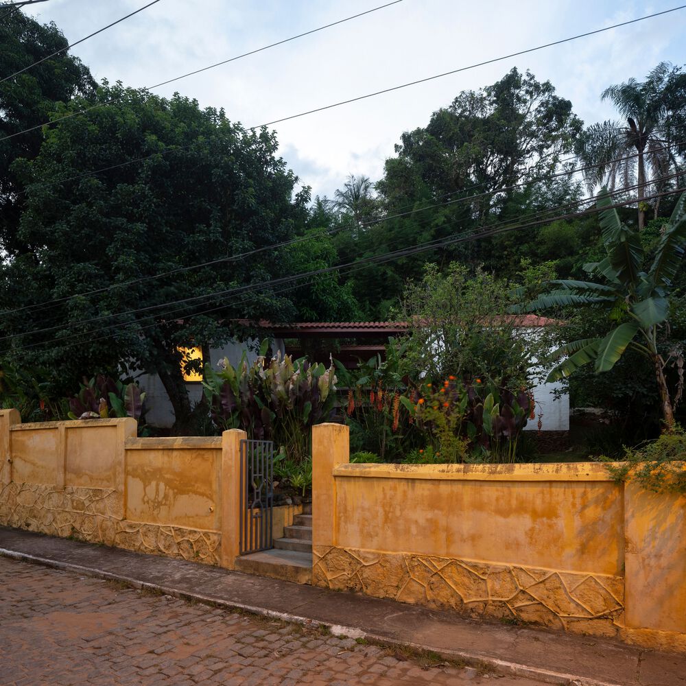 Vista da rua para a casa com muro de cerca de um metro de altura em tom amarelo envelhecido, com jardim tropical e casa ao fundo.