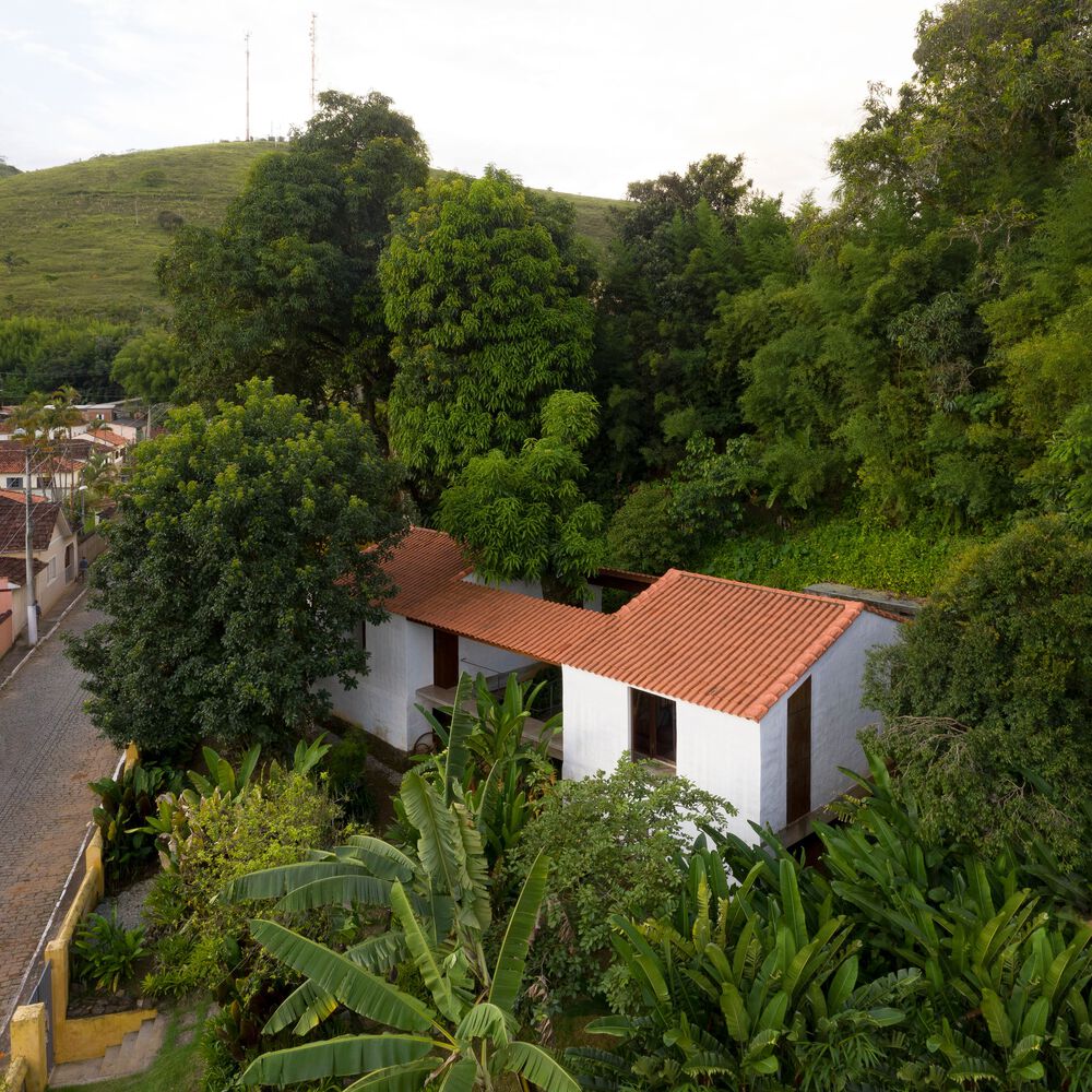 Vista aérea mostra casa em dois volumes conectados por pátio central com grande árvore, em meio ao terreno com vegetação tropical densa.