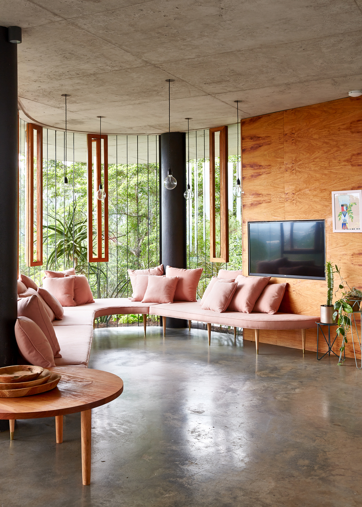 Sala de estar e TV com sofá cor de rosa e panos de vidro que permitem vista para a floresta.